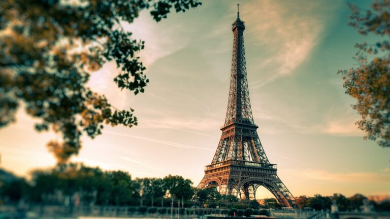 A Tall Metal Tower With Trees In The Background With Eiffel Tower In The Background